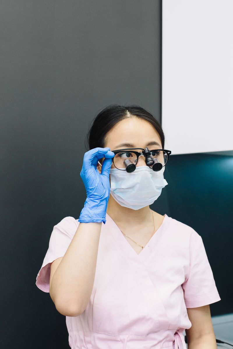 Female dentist in scrubs wearing face mask and magnifying glasses in a clinic setting.